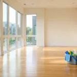 Move-out Cleaning Guide - Cleaning Tips Today A bright, empty apartment living room with sunlight reflecting off clean hardwood floors and a bucket of cleaning supplies in the corner.