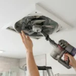 Hands cleaning bathroom exhaust fan blades with a vacuum brush attachment