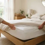 Two people rotating a made bed mattress in a sunlit bedroom with neutral tones