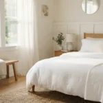 A neatly made bed with clean white sheets and a folded blanket in a bright, airy bedroom with morning light.