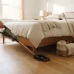 Person using a slim vacuum to clean the hardwood floor under a neatly made bed in a bright bedroom
